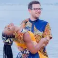 Bride and groom Alex Miller on a sandy beach; Demola holds Uche as she leans back. Both wear matching blue, yellow, brown outfits. Water glistens.
