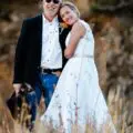 James, wearing a black jacket and jeans, stands beside Emily in a white dress, outdoors on grass with trees and rocks behind them.