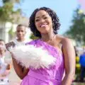 Annie in a bright pink dress walks down an outdoor red carpet, smiling and holding a white fan, with people and flowers behind.