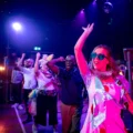 A group of people dance enthusiastically on a stage under colorful lights, with Sarah in the foreground wearing heart-shaped sunglasses and a polka dot dress.