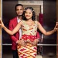 Demola stands behind Uche in an open elevator; Uche wears a colorful patterned dress, Demola a red blazer. Both smile at the camera.