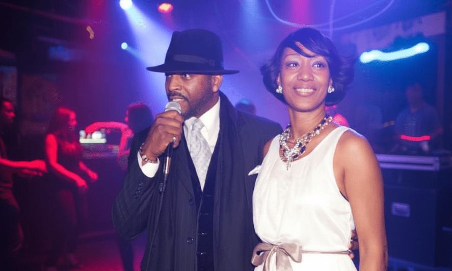 A top NYC MC Euneek of Maestro Beats DJ & Entertainment Service - in a suit and hat speaks into a mic beside a celebrant, a Mastrobeats DJ Service corporate client in a white dress